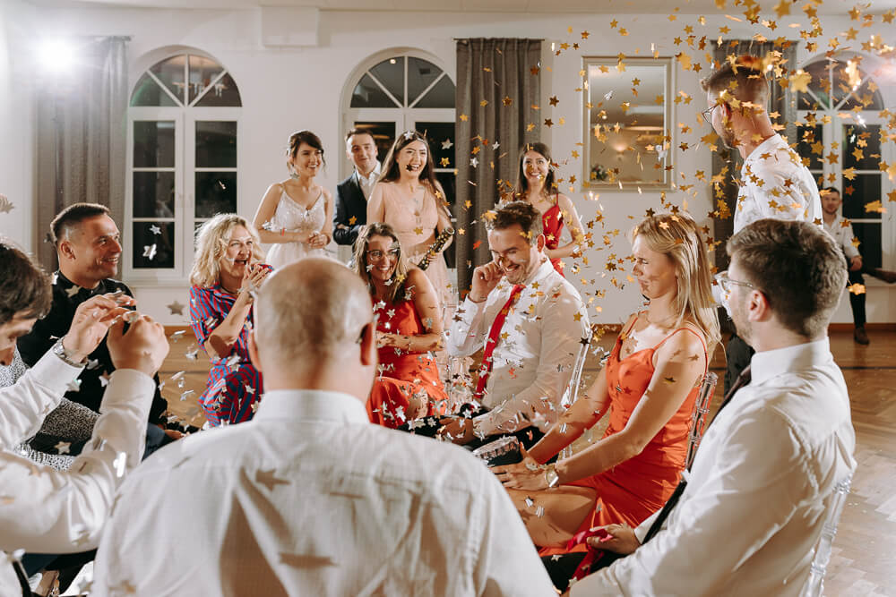 Reportaż ślubny z wesela zabawa weselna radośni goście weselni siedzą w kółku na krzesełkach confetti złote gwiazdy