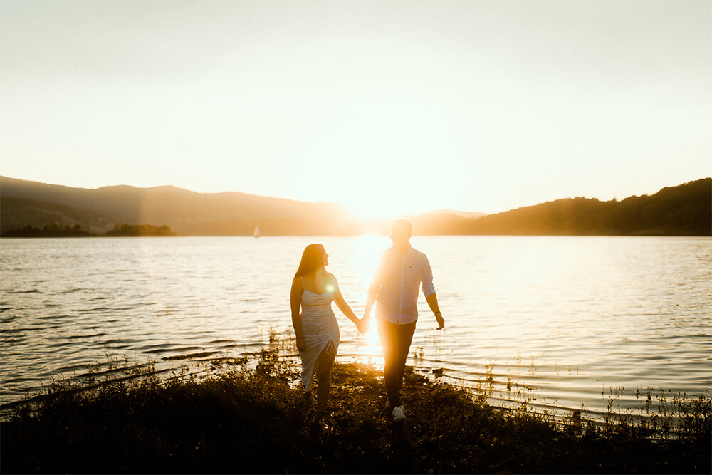 Sesja narzeczeńska w plenerze nad Jeziorem Mucharskim narzeczeni idą razem i trzymają się za rękę przy zachodzącym słońcu