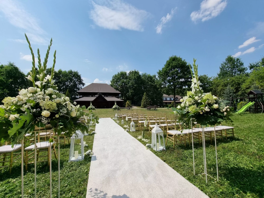 Dekoracja ślubna w Folwarku Zalesie. Biały dywan, kwiaty, krzesła dla gości i drewniana sala weselna w tle.