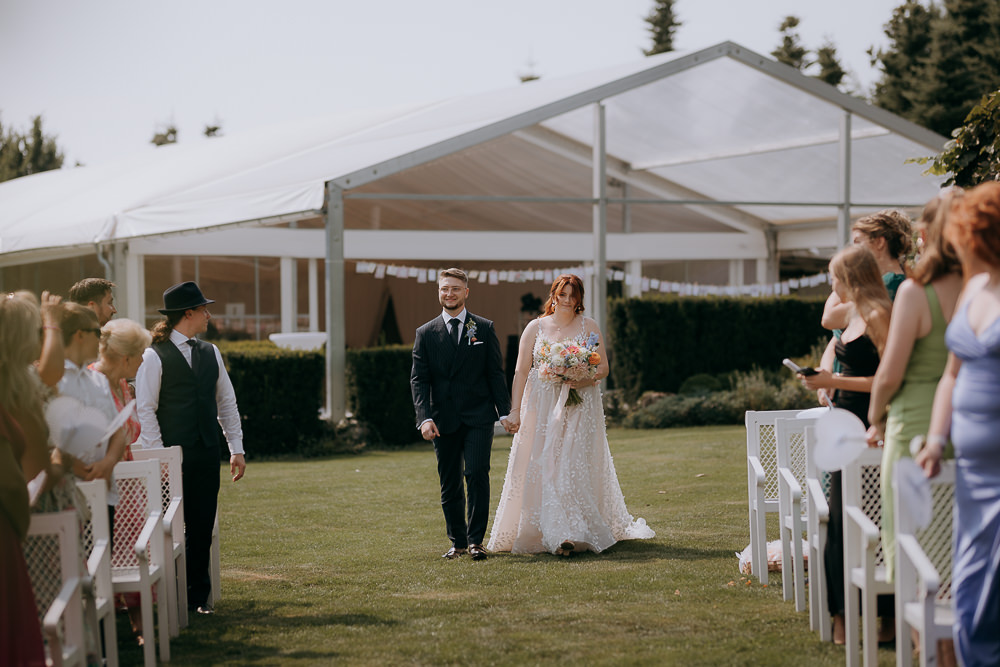 Para młoda idzie razem w plenerze, w tle biały namiot i goście. Romantyczna ceremonia ślubna.
