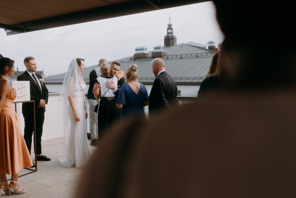 Goście gratulują młodej parze po ceremonii ślubnej, w tle budynki Krakowa.