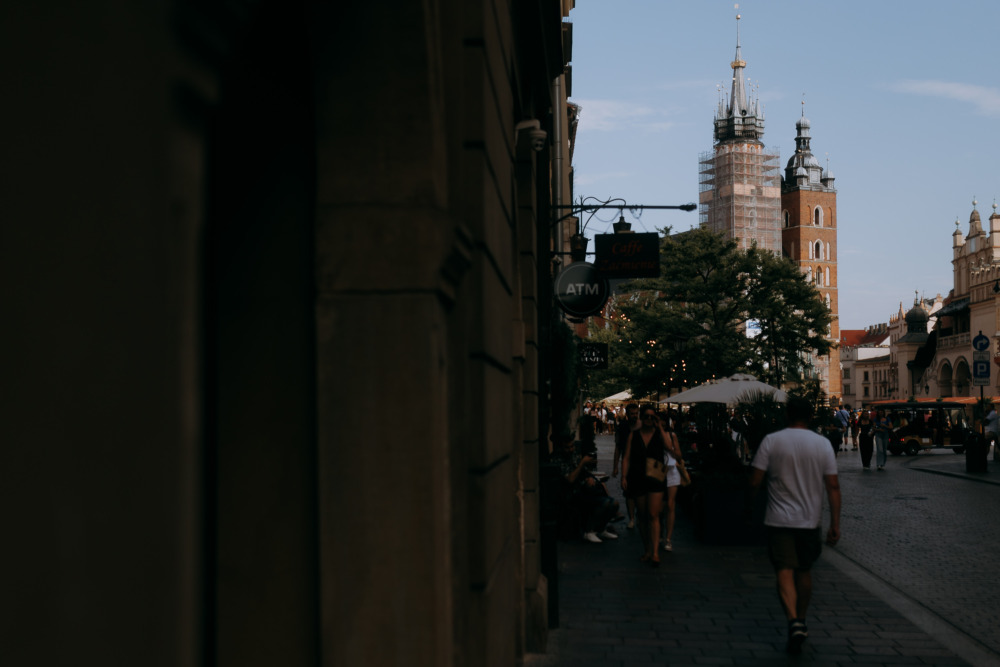 Ulica w Krakowie z Bazyliką Mariacką w tle i przechodzącymi ludźmi.