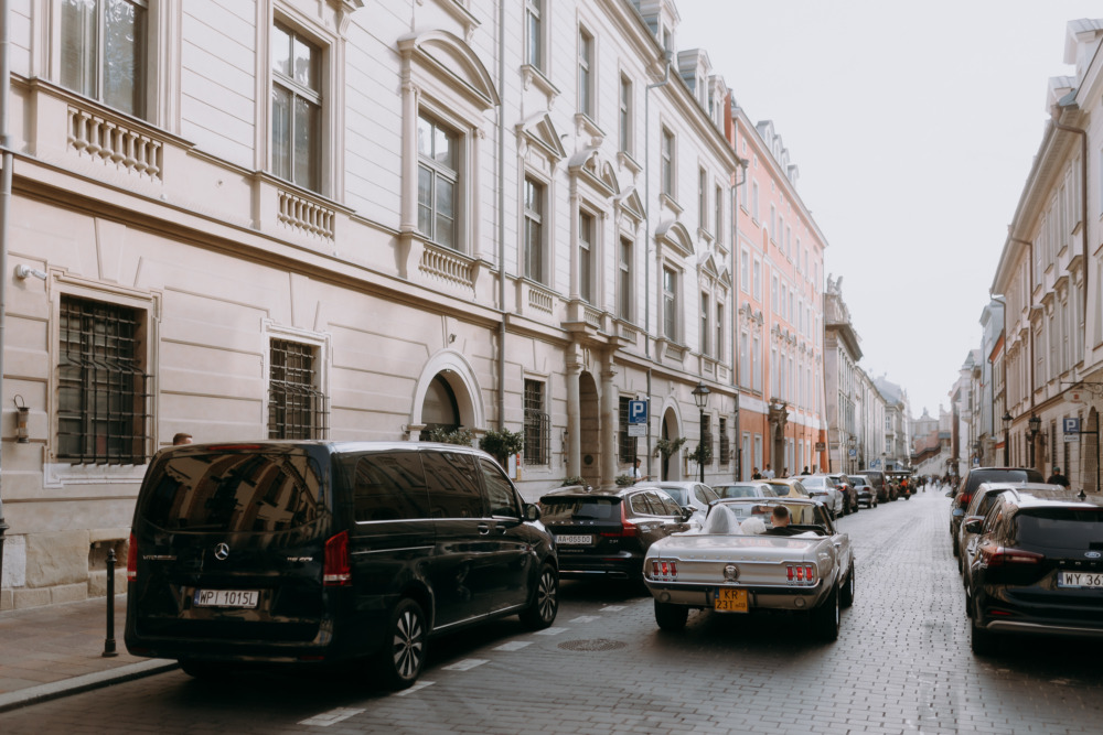 Ulica w Krakowie z zaparkowanymi samochodami, w tym Mercedes V-klasa i zabytkowy kabriolet Ford Mustang.