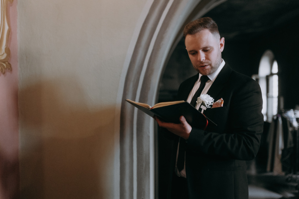 Elegancki mężczyzna czyta książkę w kościele, ubrany w czarny garnitur i krawat.