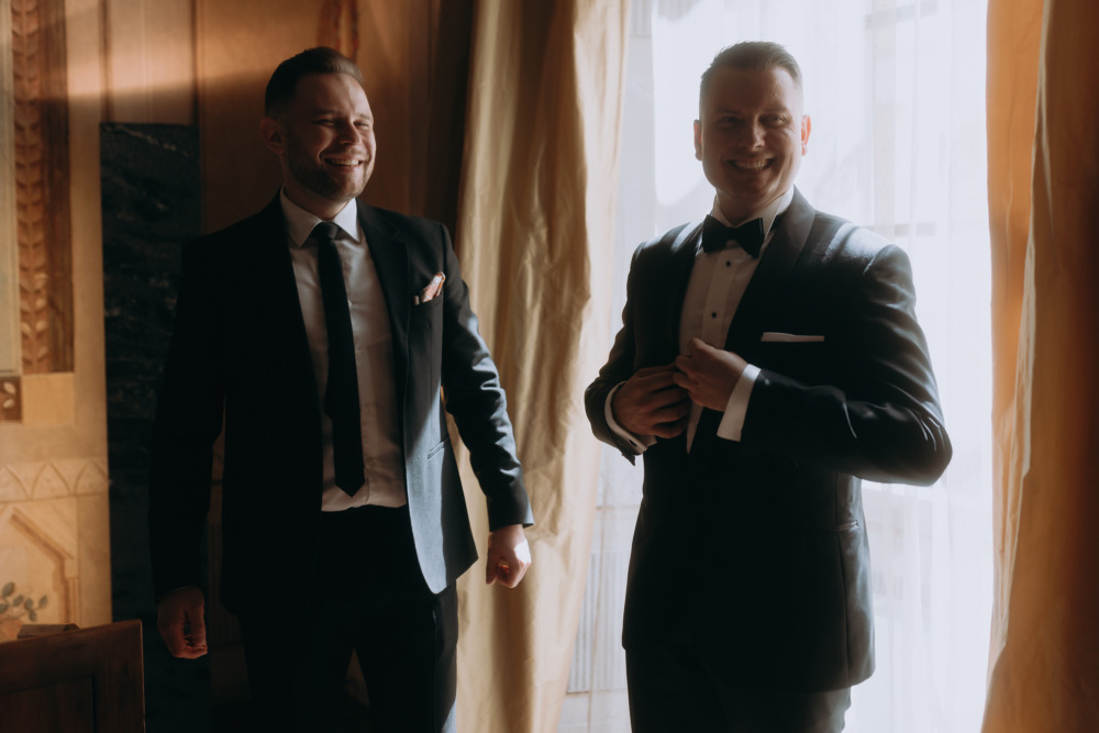 Uśmiechnięci panowie w eleganckich garniturach, przygotowania do ślubu, światło okienne.