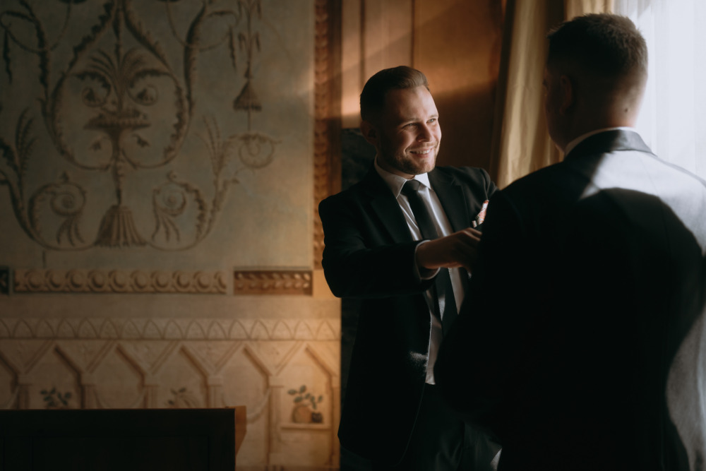 Uśmiechnięty pan młody poprawia marynarkę świadkowi w eleganckim wnętrzu.