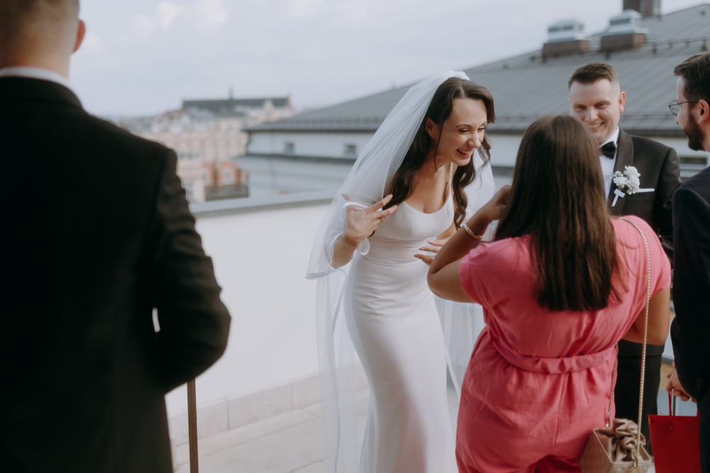 Uśmiechnięta panna młoda w sukni ślubnej rozmawia z gośćmi na tle Krakowa.