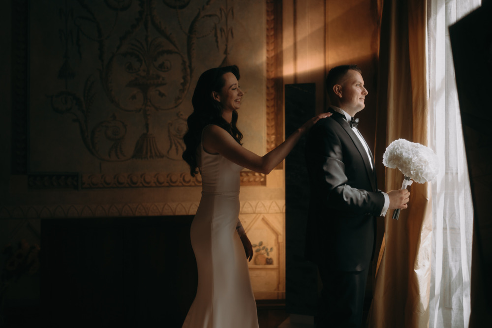 Panna młoda dotyka ramienia pana młodego z bukietem, eleganckie wnętrze