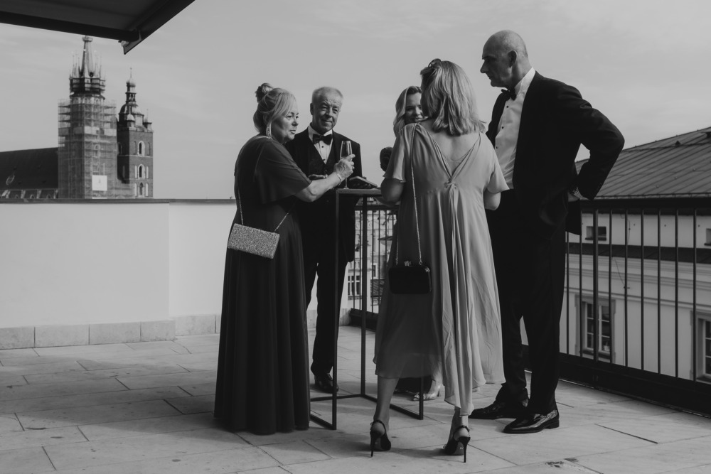 Eleganccy goście weselni w Krakowie rozmawiają na tarasie, w tle Bazylika Mariacka. Czarno-biała fotografia.