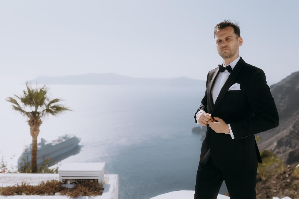 Elegancki pan młody w smokingu na Santorini, z widokiem na morze i palmę.