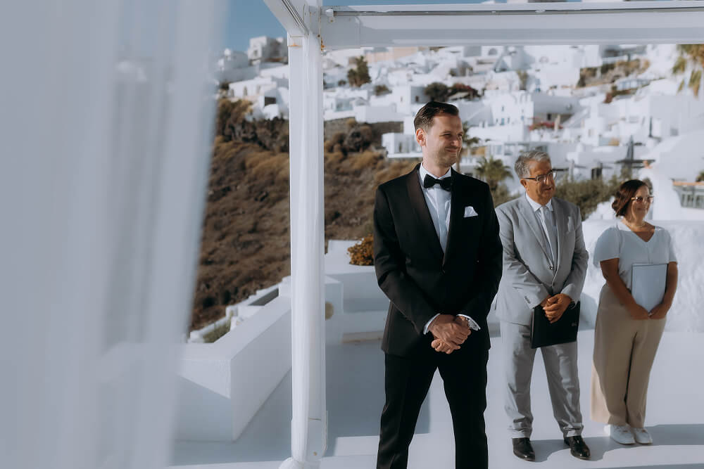 Pan młody czeka na ślub na Santorini, w tle białe budynki i urzędnicy.