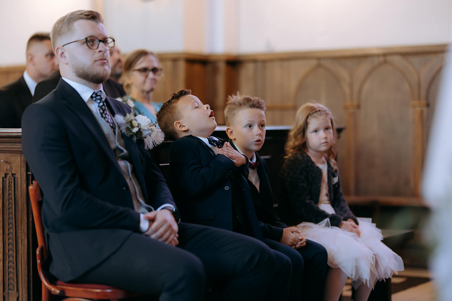 Goście weselni siedzą w ławkach podczas ceremonii ślubnej, jeden z chłopców śpiewa.