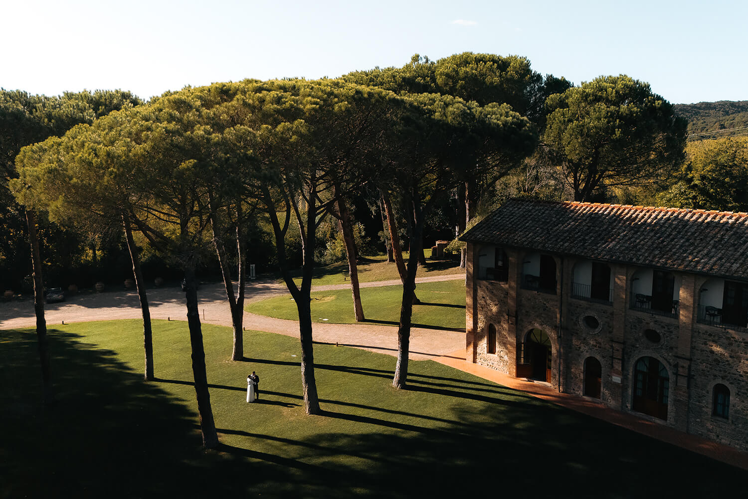 Para młoda w Toskanii spaceruje po zielonym trawniku w cieniu drzew. Kamienna budowla w tle.